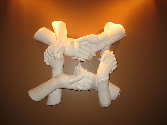 An image of four sculpted hands in white plaster interlocked and holding one another, with a soft yellow light glowing behind them.